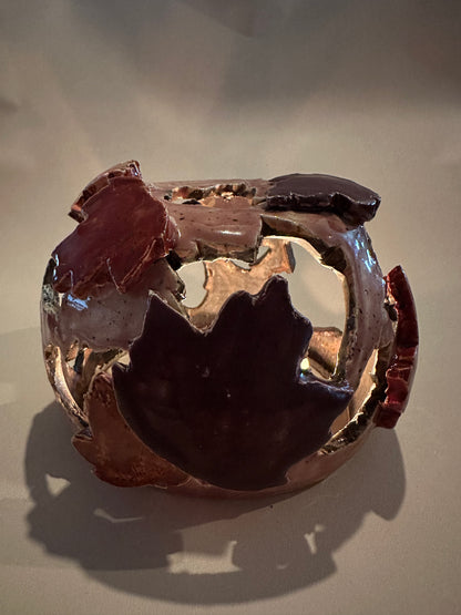 A decorative ceramic sphere with glossy mauve base glaze, featuring maple leaf-shaped cutouts in various shades of red, with three lit tea lights inside, on a neutral beige background.