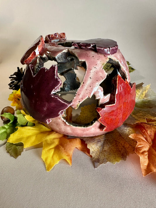 A decorative ceramic sphere with glossy mauve base glaze, featuring maple leaf-shaped cutouts in various shades of red, styled on artificial autumn leaves and a pinecone against a neutral beige background.