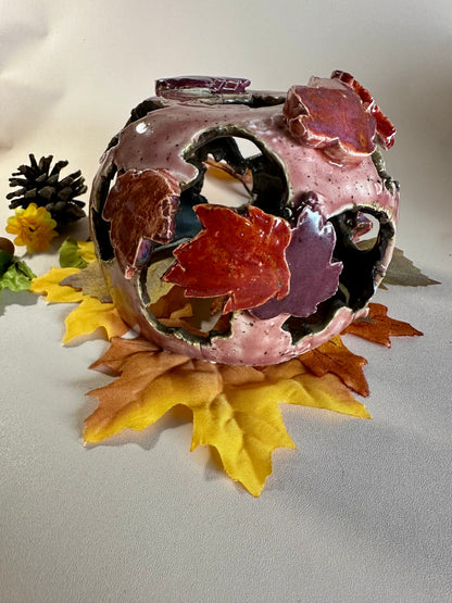 A decorative ceramic sphere with glossy mauve base glaze, featuring maple leaf-shaped cutouts in various shades of red, styled on artificial autumn leaves and a pinecone against a neutral beige background.