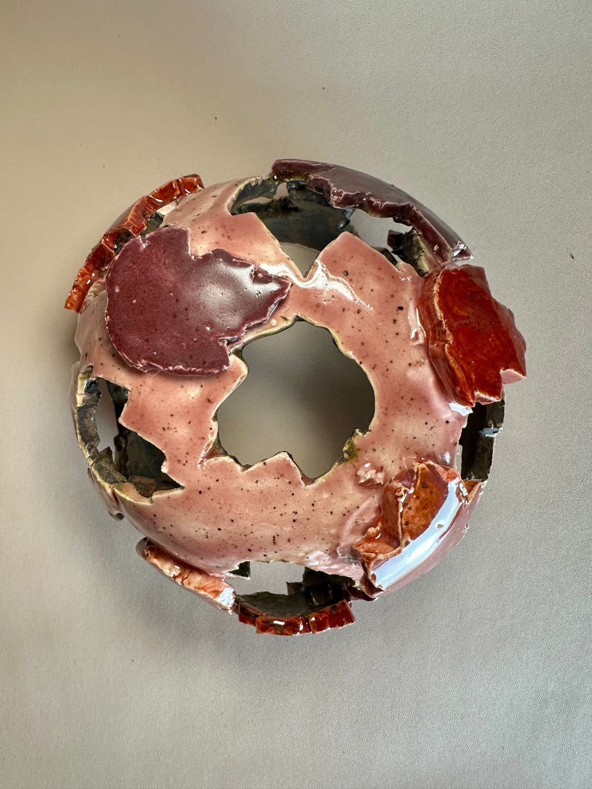A decorative ceramic sphere with glossy mauve base glaze, featuring maple leaf-shaped cutouts in various shades of red, on a neutral beige background.