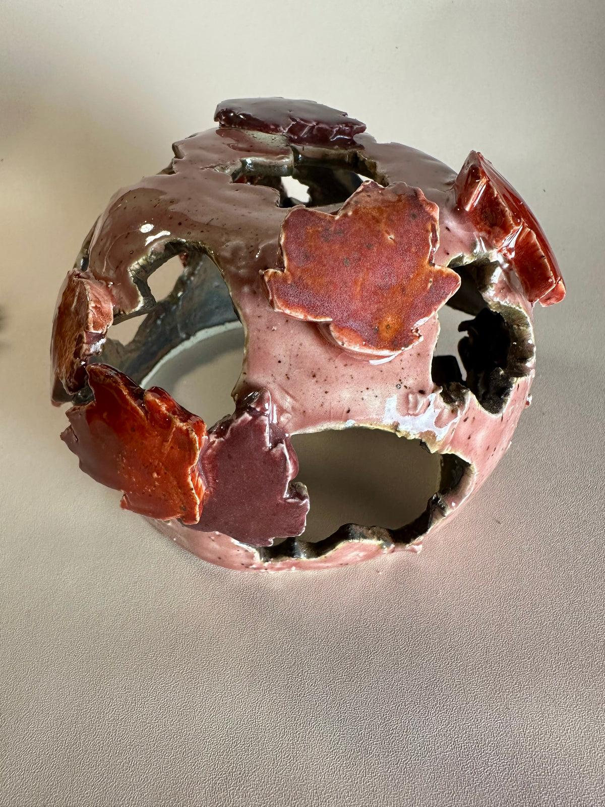 A decorative ceramic sphere with glossy mauve base glaze, featuring maple leaf-shaped cutouts in various shades of red, on a neutral beige background.