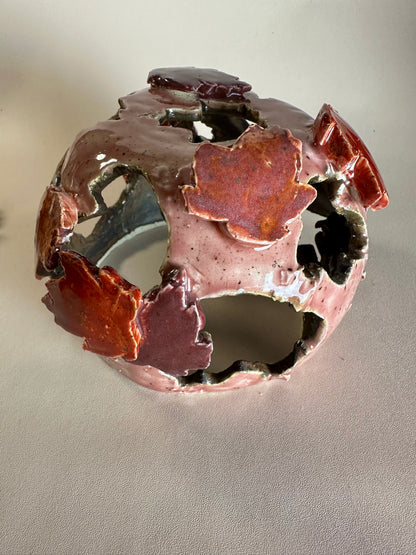 A decorative ceramic sphere with glossy mauve base glaze, featuring maple leaf-shaped cutouts in various shades of red, on a neutral beige background.