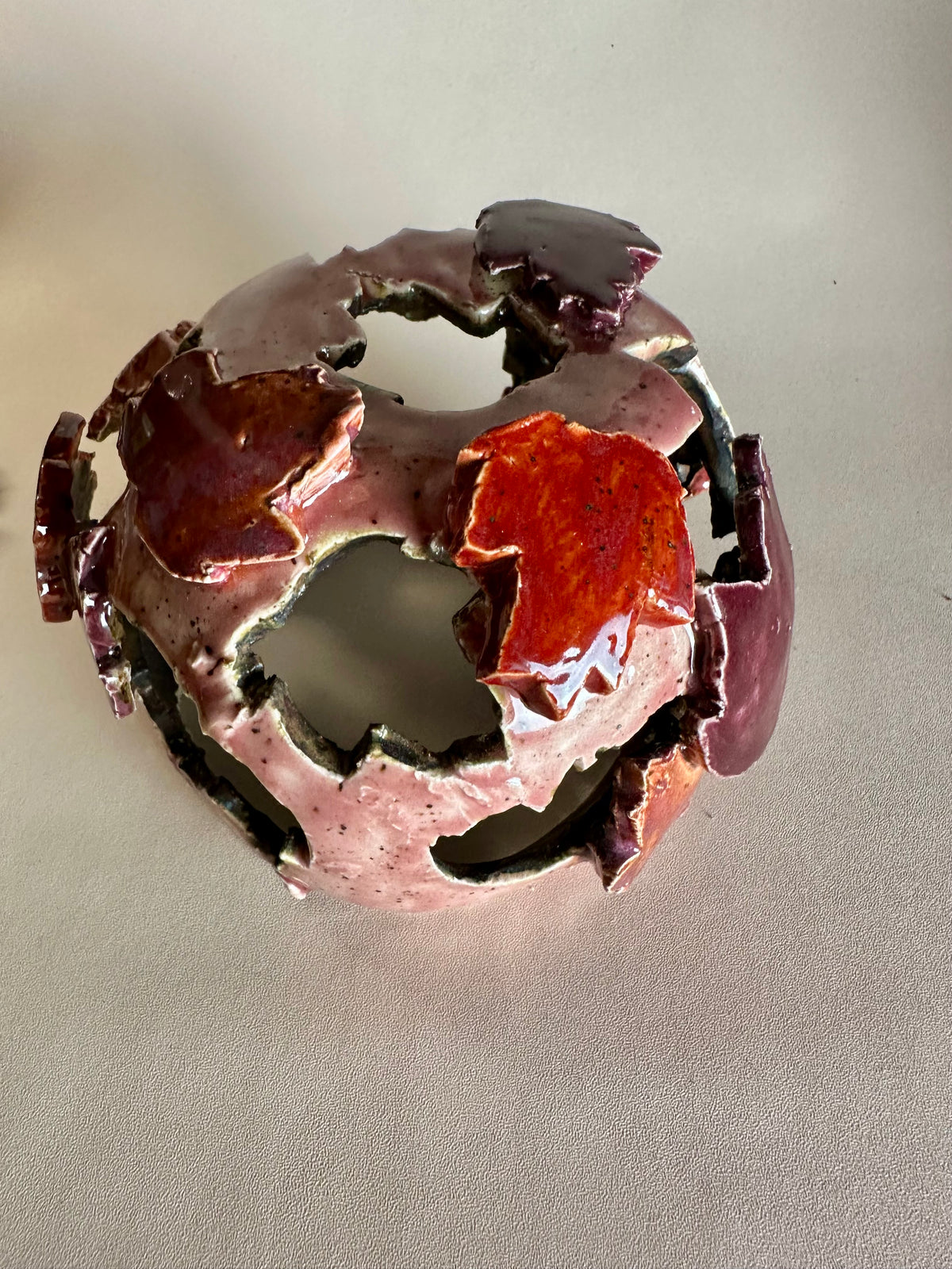 A decorative ceramic sphere with glossy mauve base glaze, featuring maple leaf-shaped cutouts in various shades of red, on a neutral beige background.