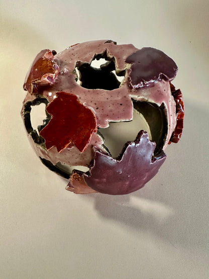 A decorative ceramic sphere with glossy mauve base glaze, featuring maple leaf-shaped cutouts in various shades of red, on a neutral beige background.