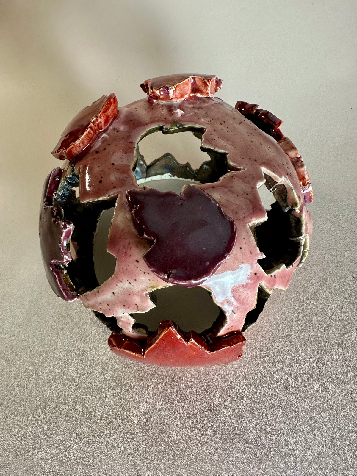 A decorative ceramic sphere with glossy mauve base glaze, featuring maple leaf-shaped cutouts in various shades of red, on a neutral beige background.