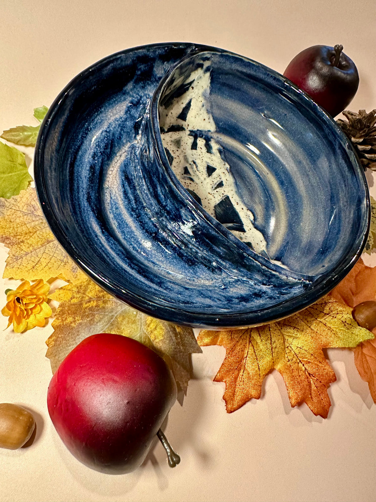 A ceramic serving bowl with a deep blue and white marbled swirls with half-spider web pattern, styled with autumn leaves, acorns, and red apples on a beige background.