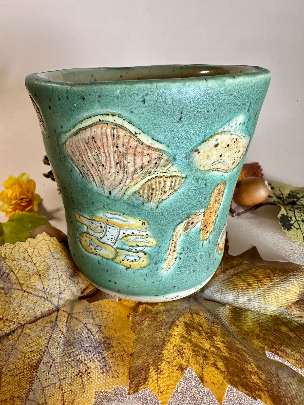 A jade green ceramic cup with brown, orange, yellow, tan, and white mushroom illustrations, stylized with fall foliage underneath on a solid neutral backdrop.