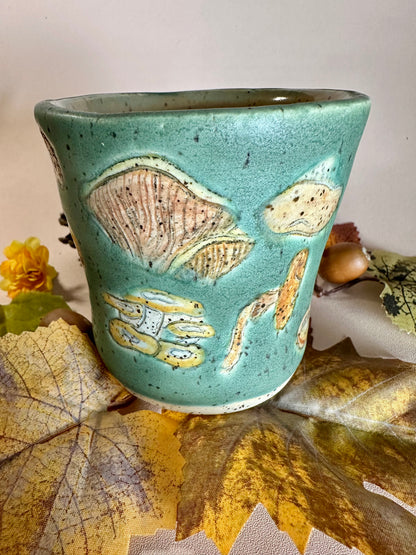 A jade green ceramic cup with brown, orange, yellow, tan, and white mushroom illustrations, stylized with fall foliage underneath on a solid neutral backdrop.