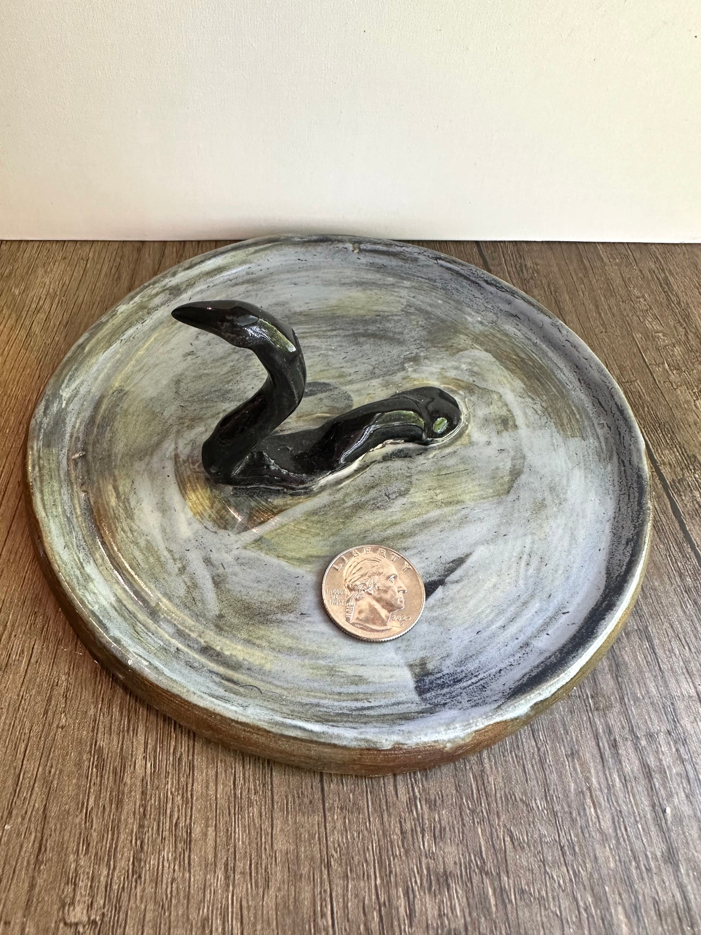 Loch Ness monster-inspired ring stand; black serpent in a pool of glossy, murky blue; coin in the blue dish for scale reference.