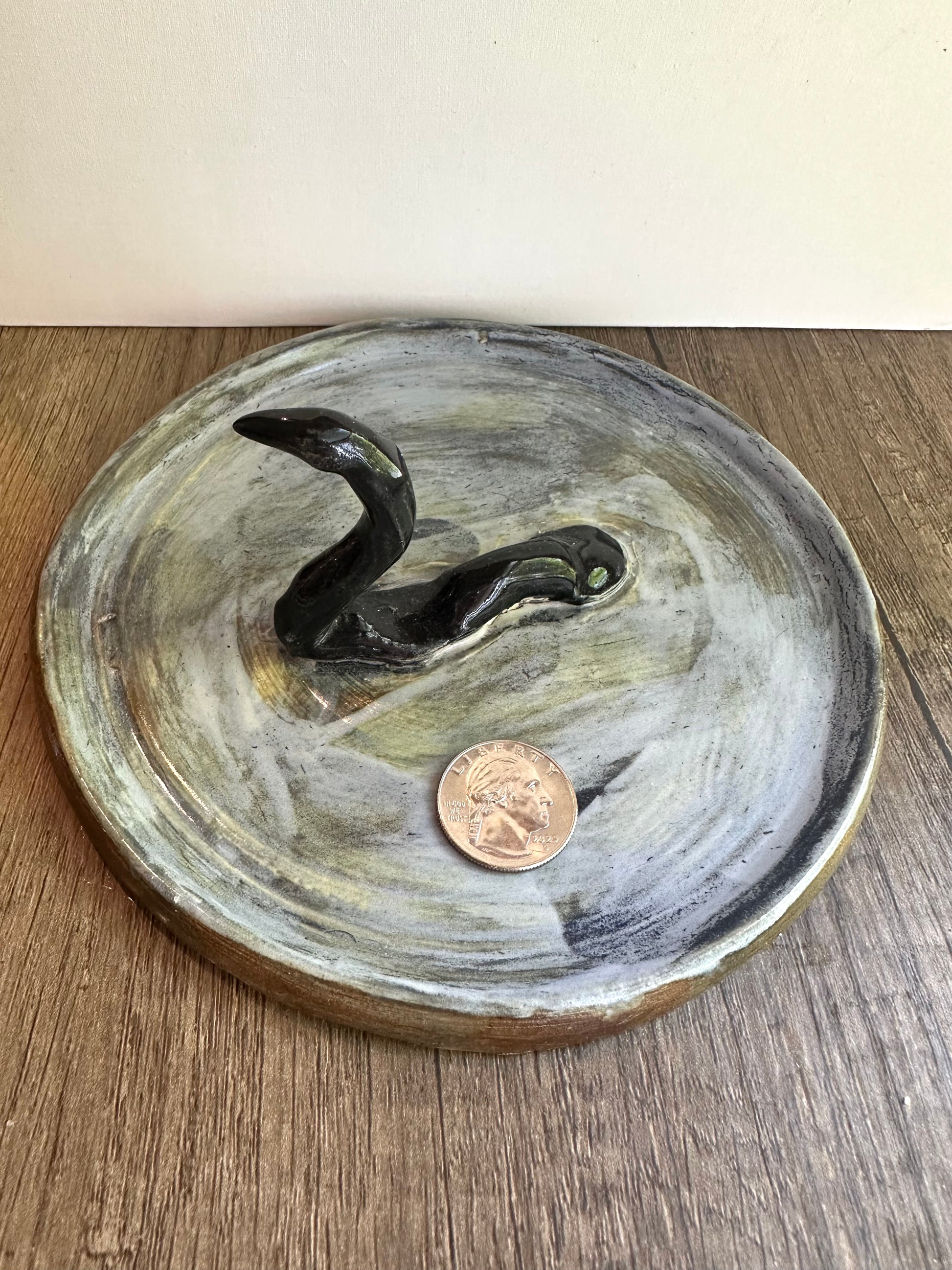 Loch Ness monster-inspired ring stand; black serpent in a pool of glossy, murky blue; coin in the blue dish for scale reference.