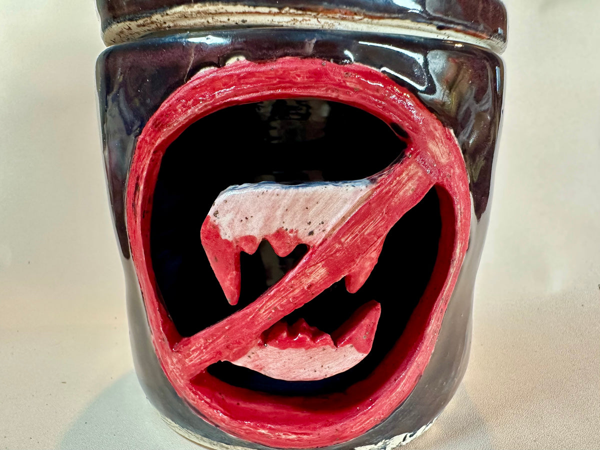 Close up of a glossy black canister with a cut out of a set of bloody vampire fangs inside a red circle with a slash. Set against a solid neutral background.