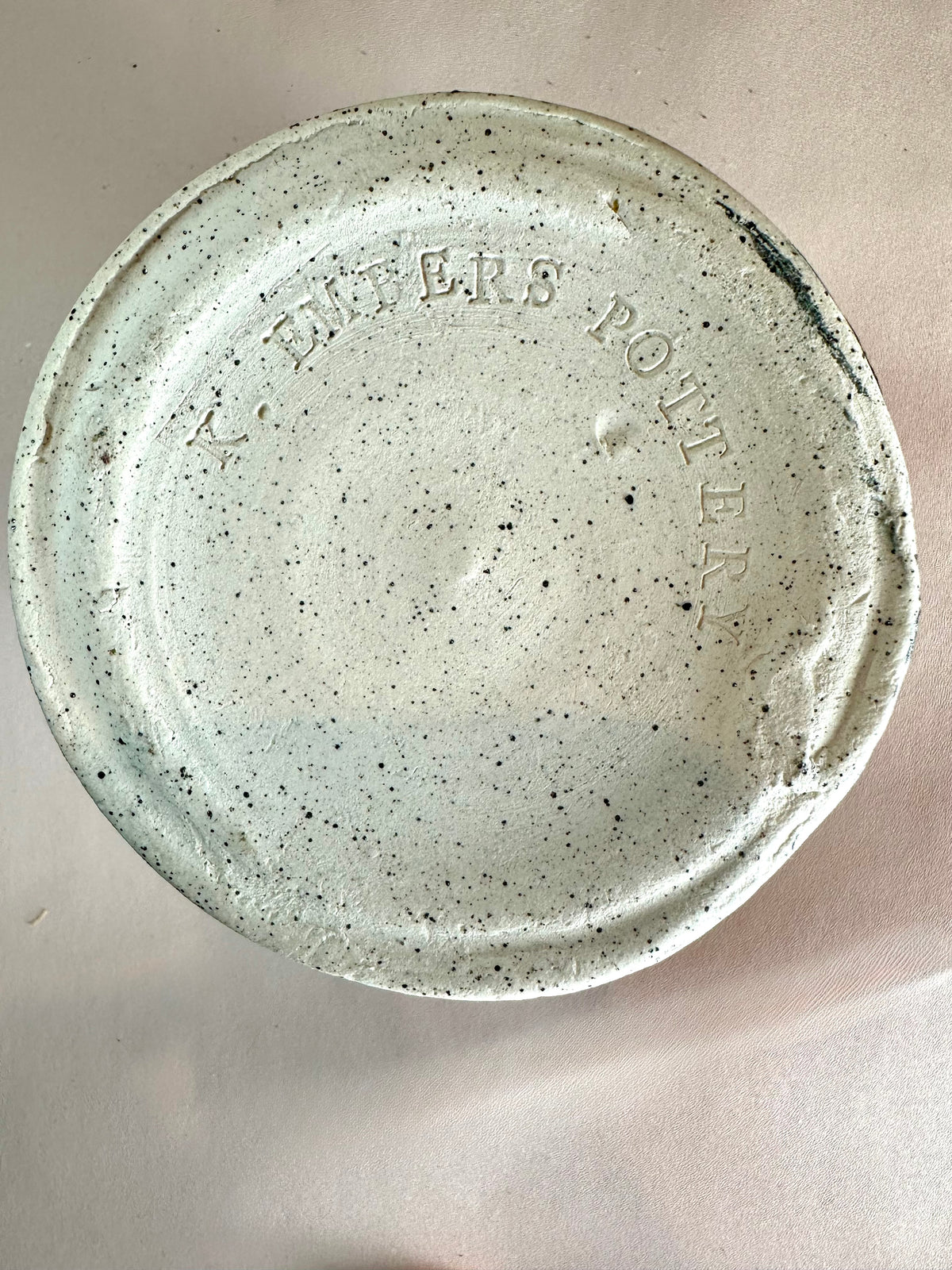 A black-speckled white clay bottom of a ceramic canister that has "K. Embers Pottery" stamped in the base.