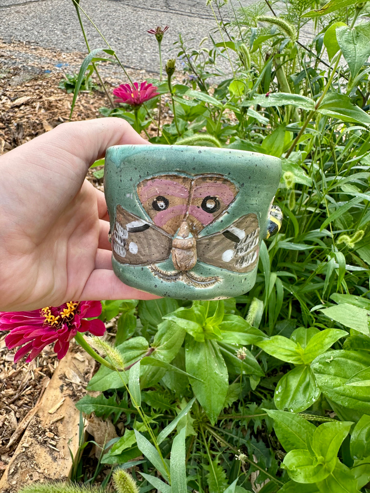 A hand holds a handmade green ceramic mug decorated with a brown Polyphemus moth; set against a garden background with a pink zinnia.