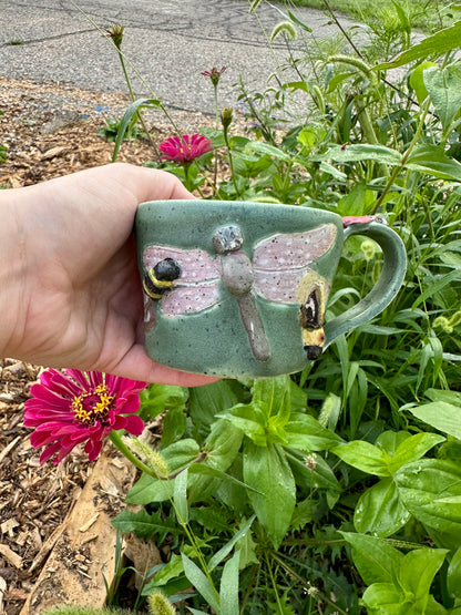 A hand holds a handmade green ceramic mug decorated with a bumblebee, purple and green dragonfly, and a firefly; set against a garden background with a pink zinnia.