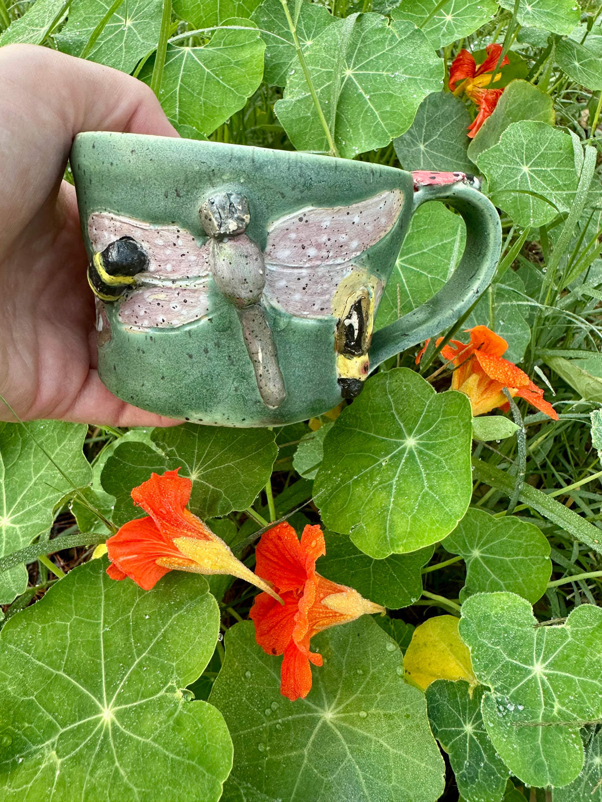 A hand holds a handmade green ceramic mug decorated with a bumblebee, purple and green dragonfly, and a firefly; set against a garden background with orange nasturtiums.