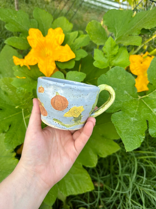 A hand holds a blue ceramic mug with illustrations of pumpkins, pumpkin flowers, and a green handle; set against a garden background of pumpkin leaves and pumpkin flowers.