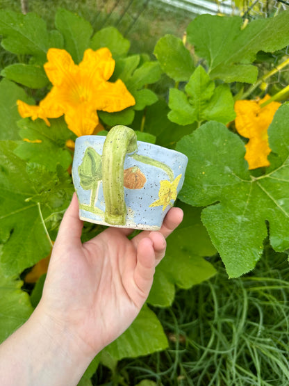 A hand holds a blue ceramic mug with illustrations of pumpkins, pumpkin flowers, pumpkin leaves, and a green handle; set against a garden background of pumpkin leaves and pumpkin flowers.