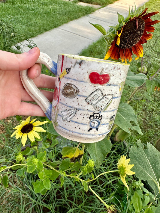 A hand holds a handmade white ceramic mug decorated as a piece of notebook paper with illustrations of books, paper airplanes, apples, pencils, analog alarm clocks, footballs, reading lamps, and to-go coffee cups. Set against a garden background with sunflowers.