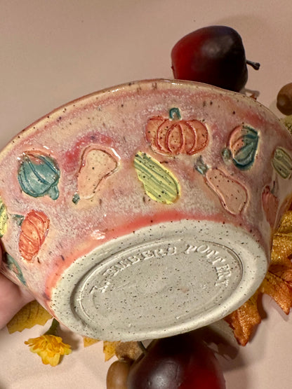 A handmade ceramic bowl with a glossy mauve glaze base, speckled off-white bottom, and a decorated exterior featuring colorful squashes and pumpkins motifs, styled with fall leaves, pinecones, acorns, and red apples on a neutral background.