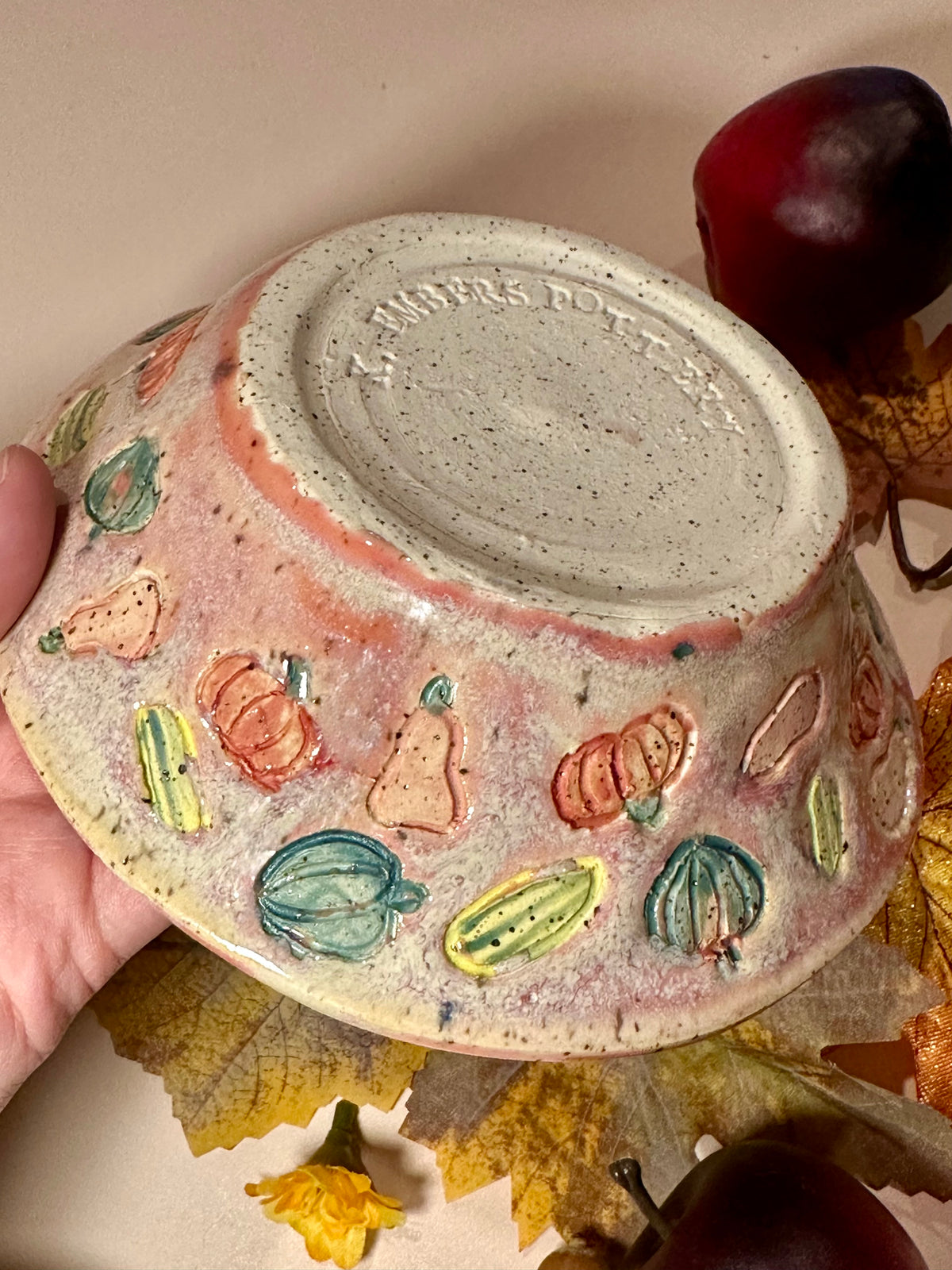 A handmade ceramic bowl with a glossy mauve glaze base, speckled off-white bottom, and a decorated exterior featuring colorful squashes and pumpkins motifs, styled with fall leaves, pinecones, acorns, and red apples on a neutral background.