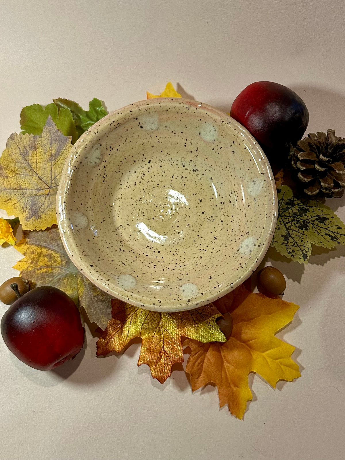 A handmade ceramic bowl with a glossy beige glaze base with dabs of a lighter off-white glaze dotting the inside just under the brim; styled with fall leaves, pinecones, acorns, and red apples on a neutral background.