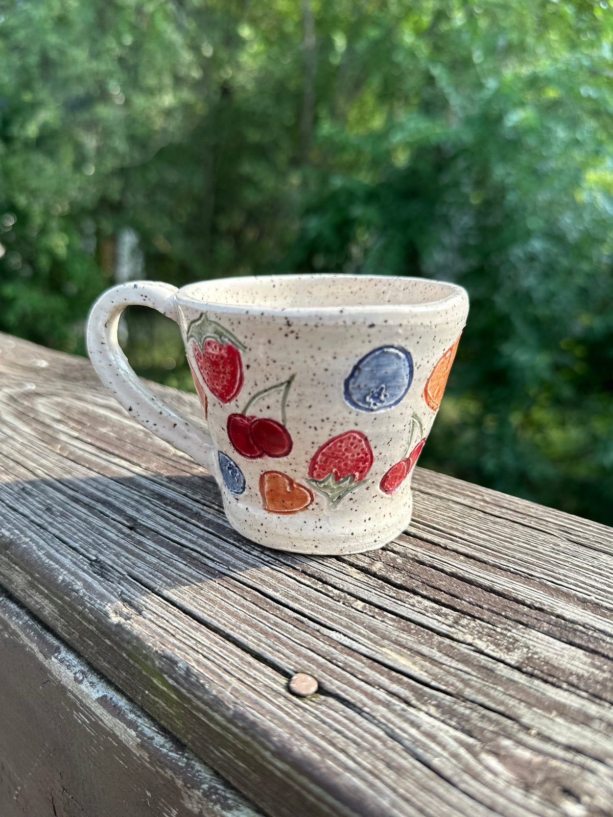 A speckled beige ceramic tea cup is decorated with painted red cherries, blueberries, strawberries and peaches, sitting on a worn wood rail set against a blurred green outdoor background.
