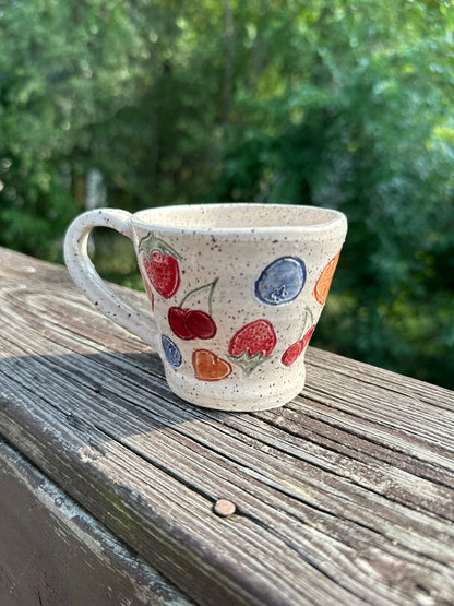 A speckled beige ceramic tea cup is decorated with painted red cherries, blueberries, strawberries and peaches, sitting on a worn wood rail set against a blurred green outdoor background.