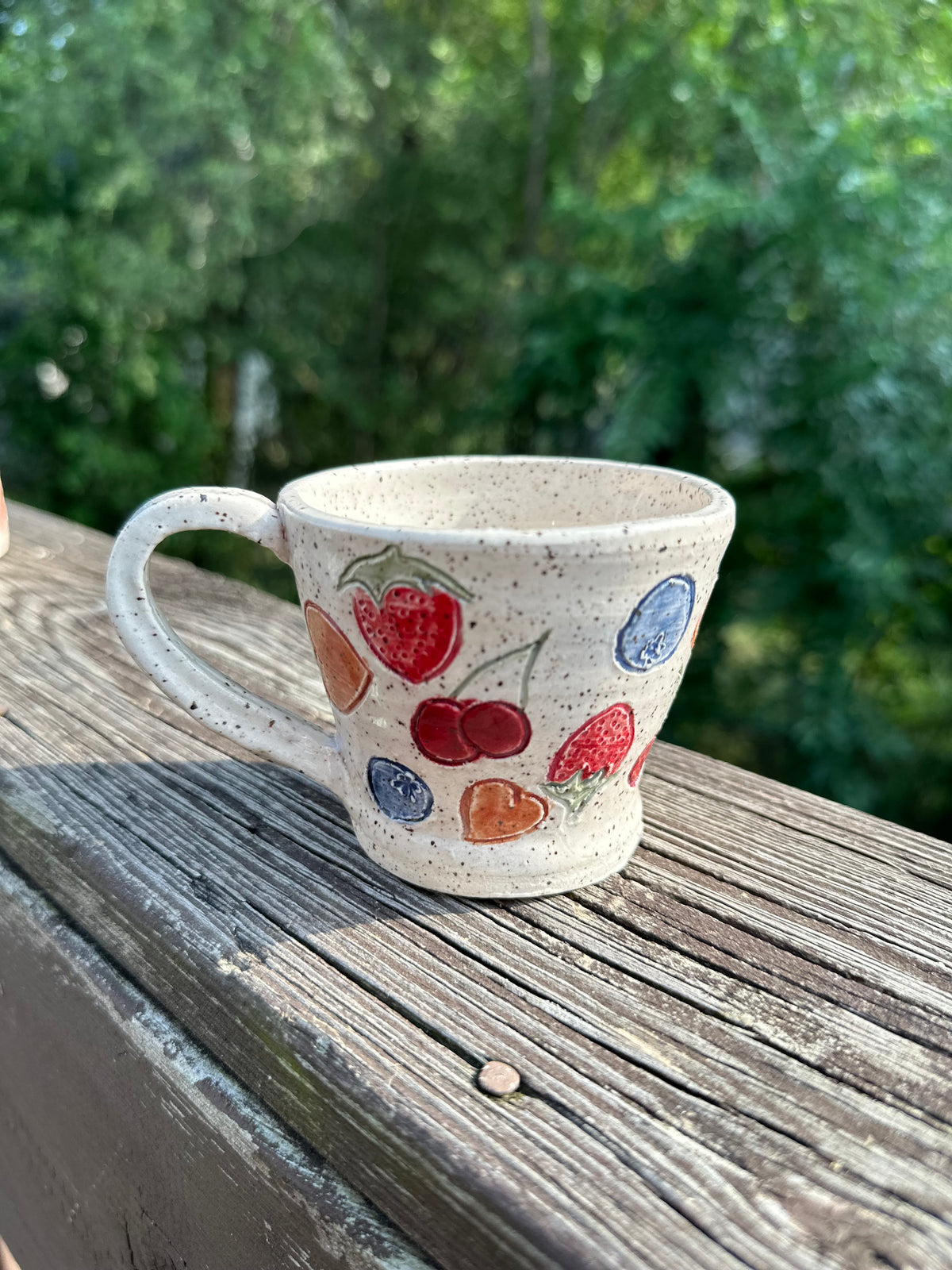 A speckled beige ceramic tea cup is decorated with painted red cherries, blueberries, strawberries and peaches, sitting on a worn wood rail set against a blurred green outdoor background.
