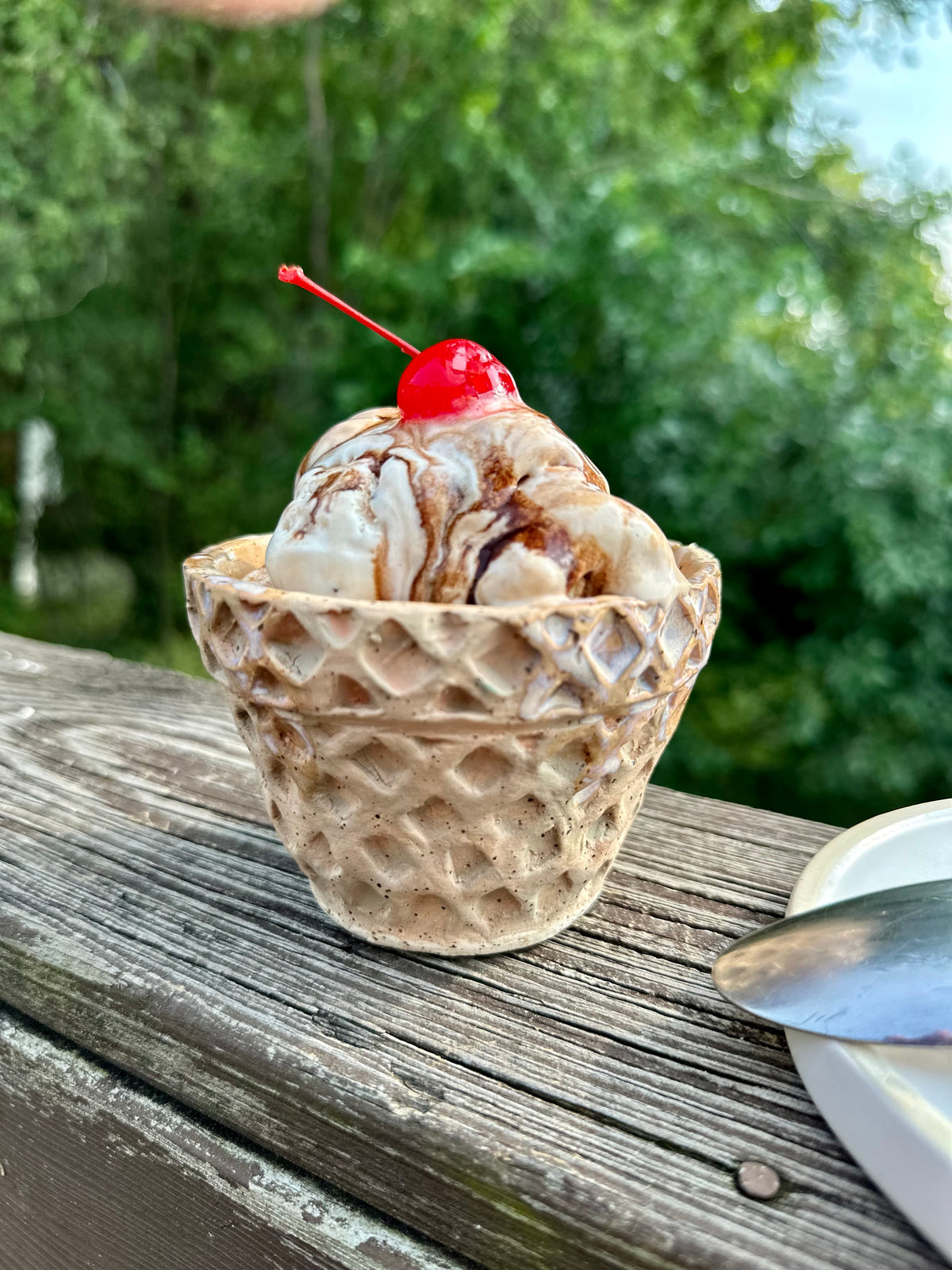 A waffle-cone-shaped dessert cup filled with vanilla ice cream and drizzled chocolate sauce, topped with a bright red cherry, next to a spoon on a rest just off to the side; sitting on a weathered wooden deck with a blurred green outdoor background.