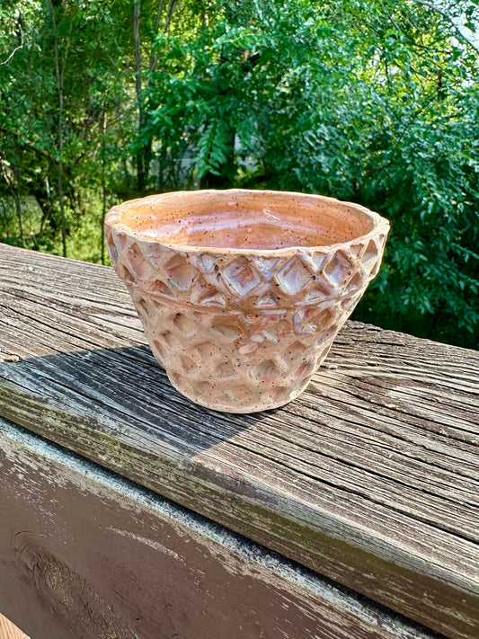 A small-medium bowl carved and colored to look like a waffle cone bowl; slight glossy glaze swirls on the brim and inside makes the illusion of dripping vanilla ice cream. Set on a worn wooden rail in front of a green forest background.